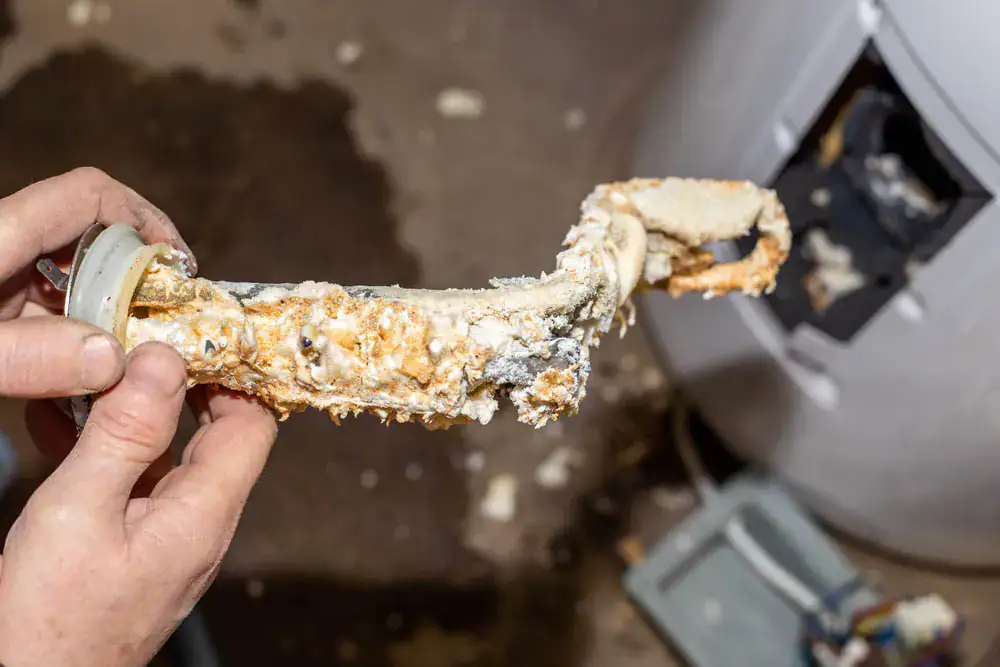 A person holds a heavily corroded and calcified heating element removed from a water heater, showing extensive mineral buildup and scale.