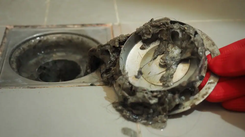 A person wearing a red glove holds a clogged shower drain cover covered in wet hair and grime, with the open drain visible on tiled flooring.