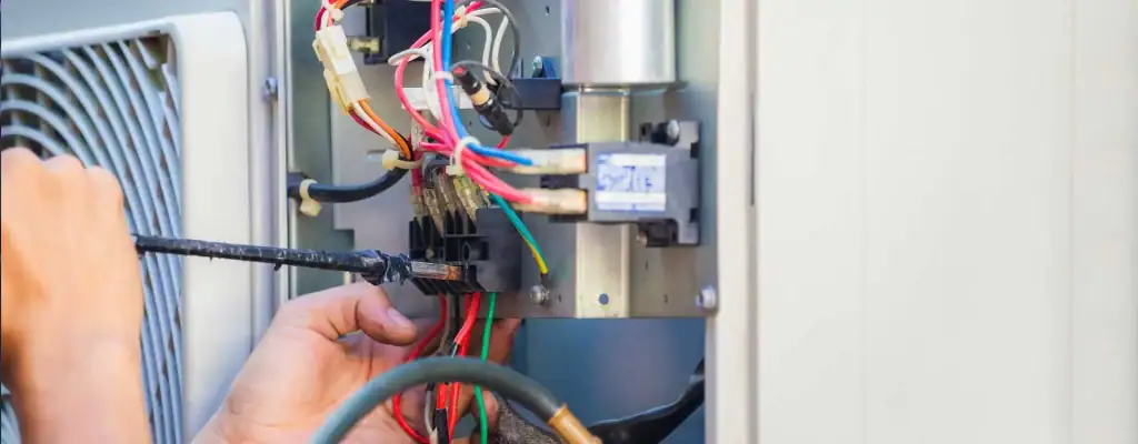 A close-up of a person’s hands repairing electrical wiring inside an HVAC unit, using a screwdriver, with colorful wires and components exposed.