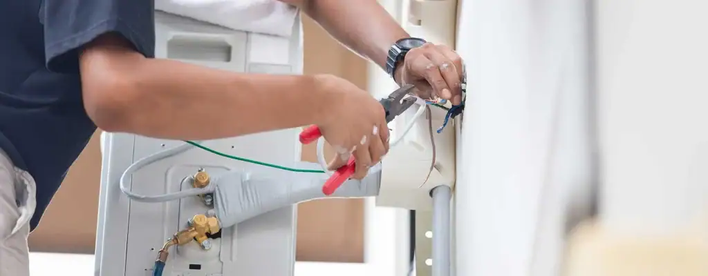 A person uses pliers to work on electrical wiring at the back of an appliance, possibly an air conditioner. Their arms and hands are visible, holding wires and tools.