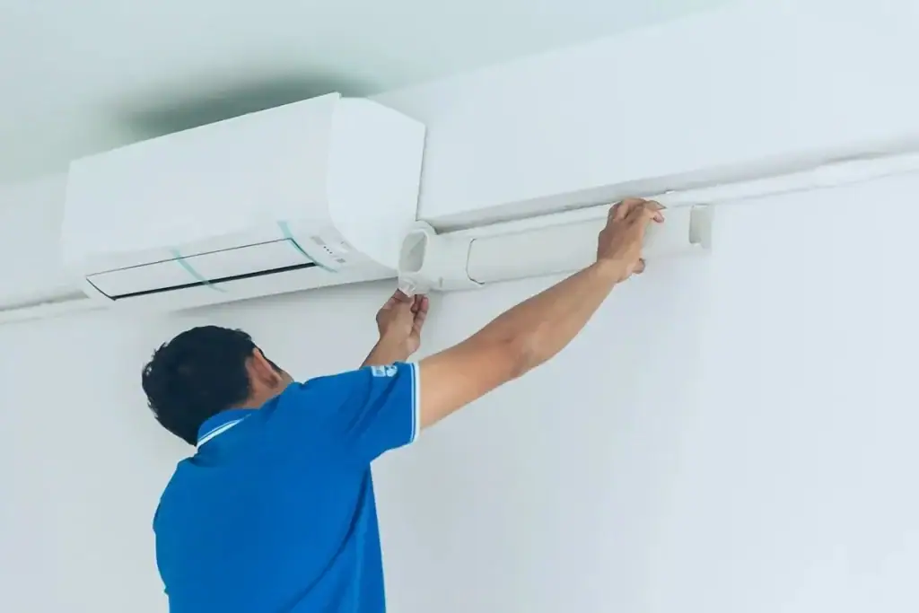 A person in a blue shirt installs or repairs an air conditioning unit mounted high on a white wall, adjusting the cover of a conduit pipe.