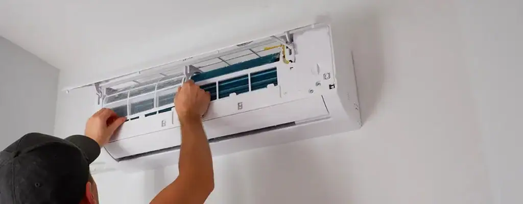 A person wearing a cap is cleaning or repairing the inside of a wall-mounted air conditioner unit with the front panel open.