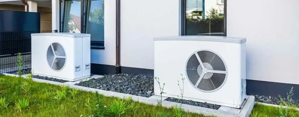 Two outdoor heat pump units are installed beside a modern building, sitting on gravel beds with some green grass and plants in the foreground.