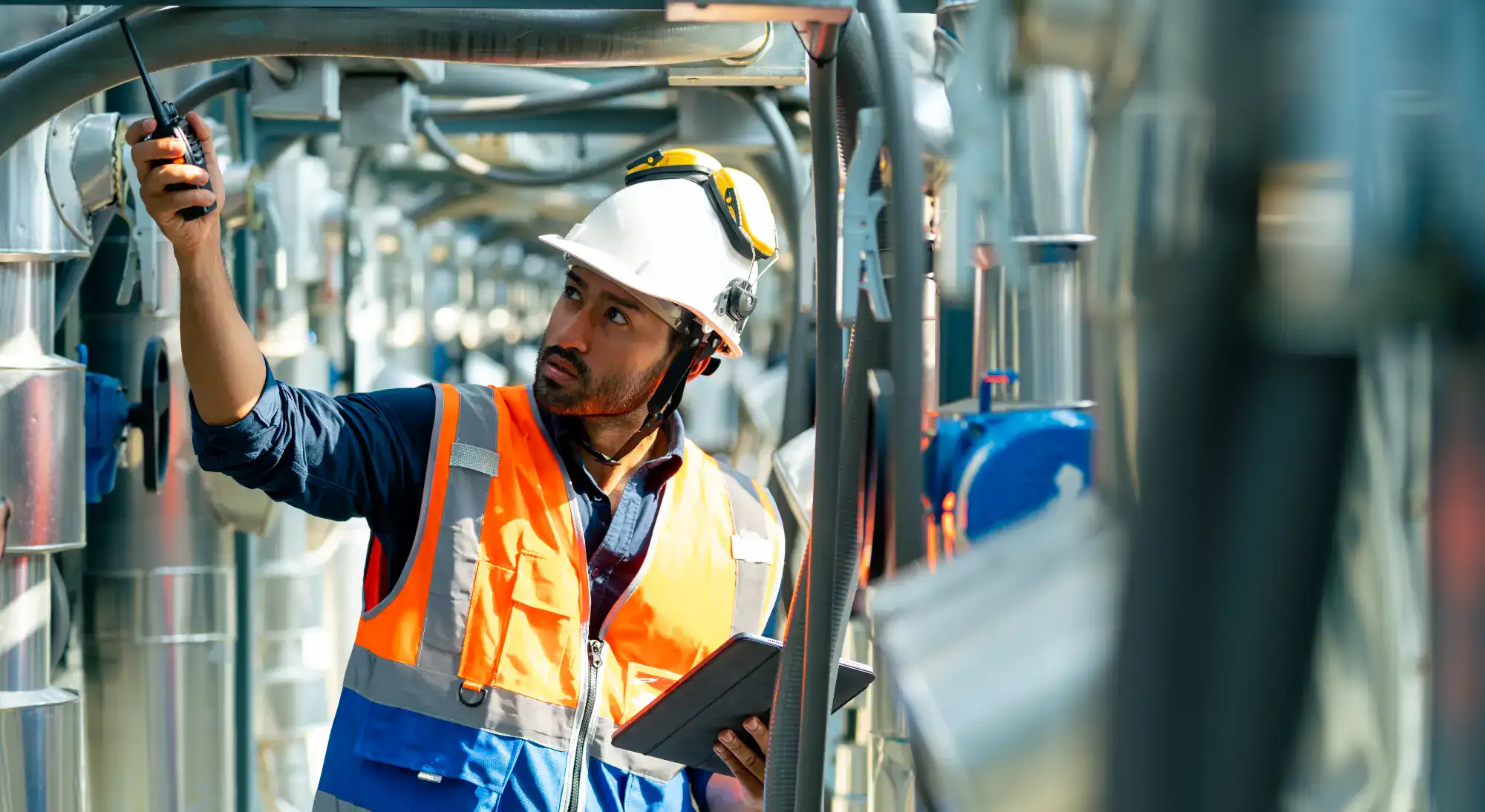 A construction worker in a white hard hat and orange safety vest holds a walkie-talkie and a tablet while inspecting industrial pipes at a worksite.