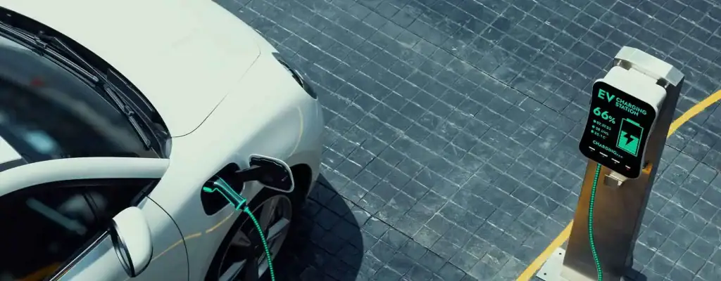 A white electric car is charging at an EV charging station on a paved surface. The charging cable is plugged into the car, and the station display shows the charging progress.