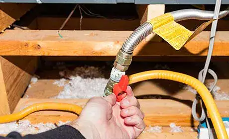A hand turns a red valve on a flexible gas pipe connection in an attic or crawl space, surrounded by wooden beams and insulation.