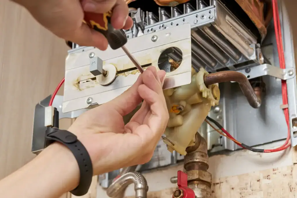 Close-up of a person’s hands using a screwdriver to repair or maintain the internal components of a gas boiler or water heater, with visible pipes and wiring.