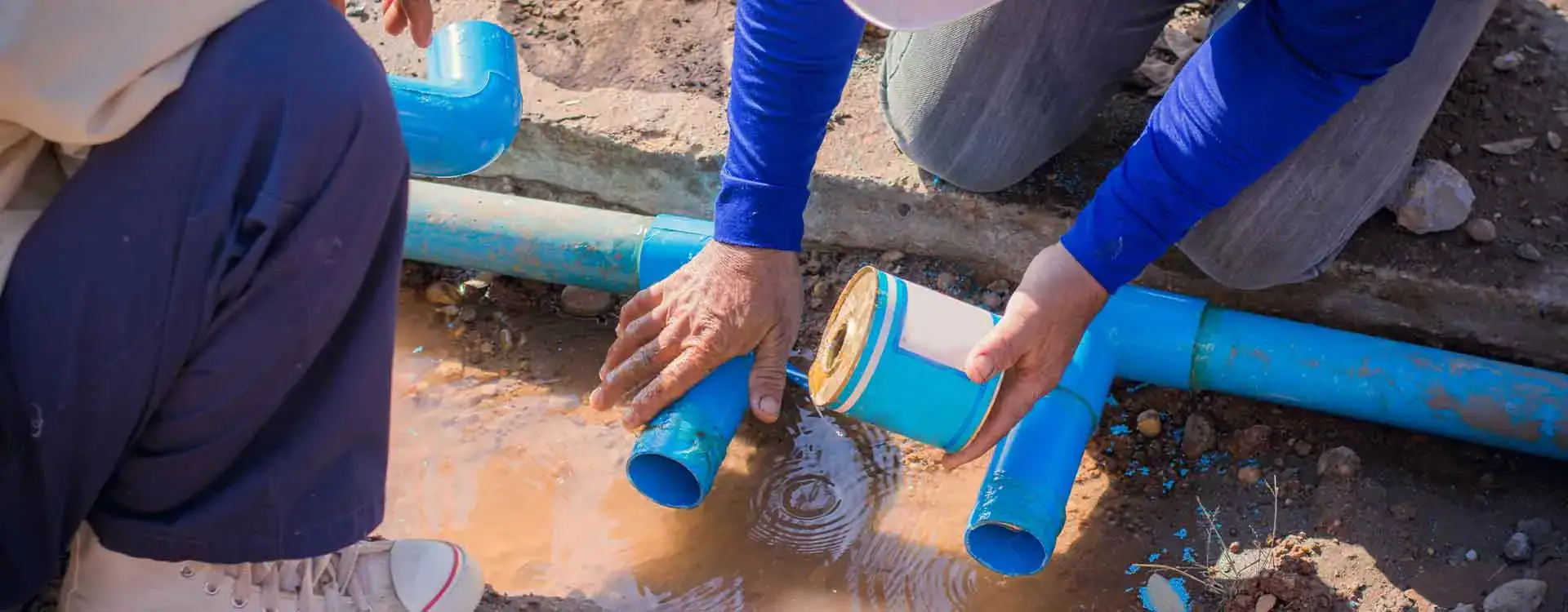 Two people are connecting blue PVC pipes on the ground near a puddle, using a can of adhesive to join the pieces for plumbing or water supply.