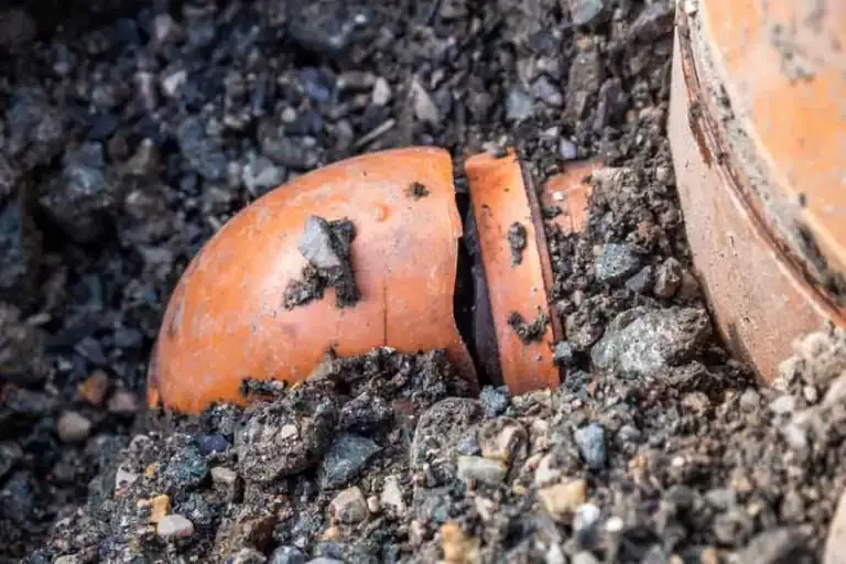 A broken orange clay pipe partially buried in gravel and dirt, with jagged edges visible where the pipe has snapped.