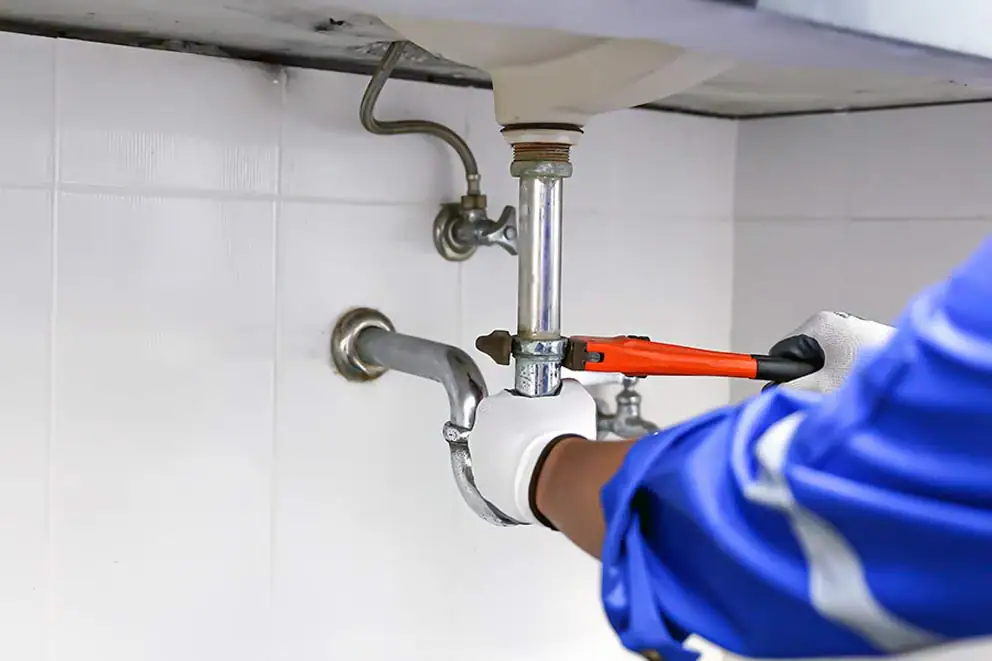 A person wearing gloves and a blue uniform is using a wrench to tighten or loosen a pipe under a sink, performing plumbing work.