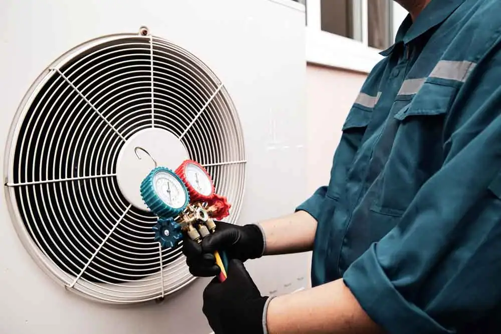 A technician in a blue uniform and black gloves checks an air conditioning unit using pressure gauges attached to the outdoor condenser unit.