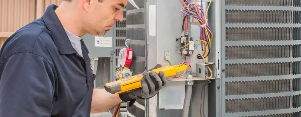 A technician wearing gloves uses a yellow multimeter to check wiring inside an outdoor air conditioning unit.