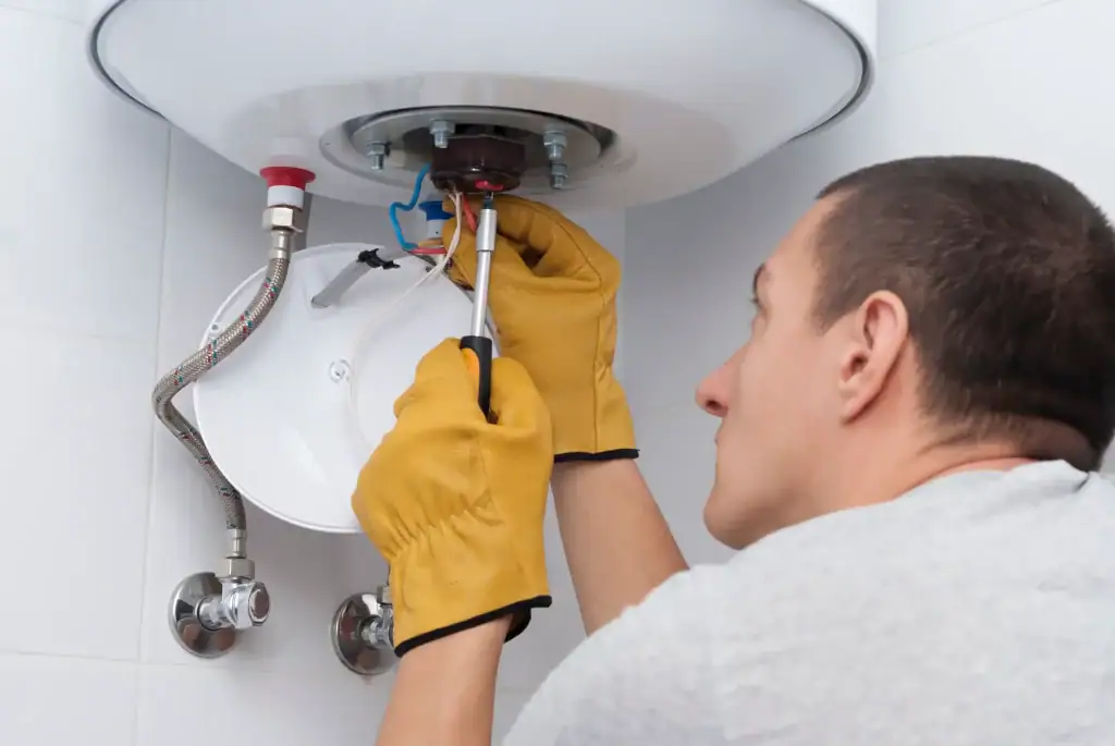 A man wearing yellow work gloves uses a screwdriver to repair a water heater mounted on a wall. Some wires and pipes are visible as he works on the heater’s underside.