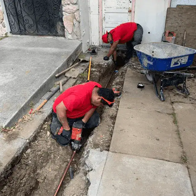 Two workers in red shirts dig a trench beside a sidewalk and house foundation, using tools to install or repair pipes. A blue wheelbarrow and various tools are nearby.